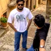 The image depicts two men in a cobblestone alleyway. One man is standing in the foreground facing the camera, wearing a white "90's Babe" t-shirt, blue jeans, and sunglasses, while another man stands to the side, wearing a dark t-shirt and jeans, with his back turned slightly toward the viewer as he leans against a stone ledge. The setting appears to be a rustic, historic street, and a logo for "The Cargo Bear" is visible in the upper right corner and at the bottom of the image