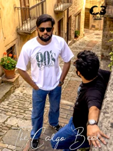 The image depicts two men in a cobblestone alleyway. One man is standing in the foreground facing the camera, wearing a white "90's Babe" t-shirt, blue jeans, and sunglasses, while another man stands to the side, wearing a dark t-shirt and jeans, with his back turned slightly toward the viewer as he leans against a stone ledge. The setting appears to be a rustic, historic street, and a logo for "The Cargo Bear" is visible in the upper right corner and at the bottom of the image