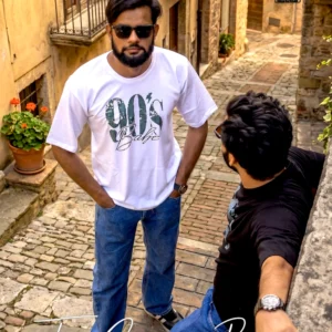 The image depicts two men in a cobblestone alleyway. One man is standing in the foreground facing the camera, wearing a white "90's Babe" t-shirt, blue jeans, and sunglasses, while another man stands to the side, wearing a dark t-shirt and jeans, with his back turned slightly toward the viewer as he leans against a stone ledge. The setting appears to be a rustic, historic street, and a logo for "The Cargo Bear" is visible in the upper right corner and at the bottom of the image